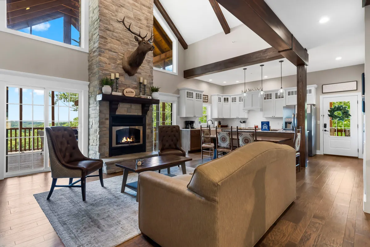 A warm, inviting living room featuring a stone fireplace, large windows that let in abundant natural light, exposed wooden beams, and a plush seating arrangement perfect for gatherings. The view out the windows highlights the scenic Ozarks beyond.