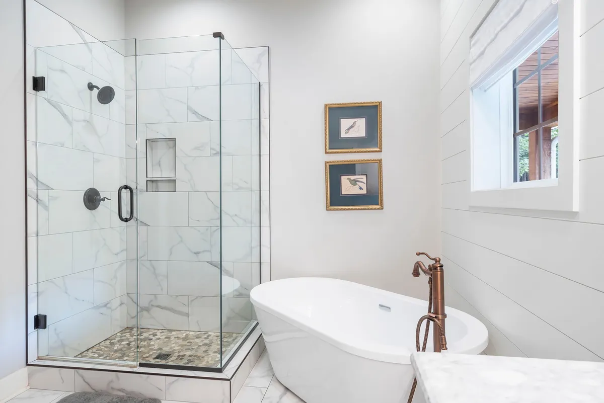 A luxurious bathroom with a glass shower enclosure, a freestanding soaker bathtub, framed artwork, and abundant natural light streaming through the window. The design blends modern comfort with relaxing touches, ideal for unwinding after a long day.