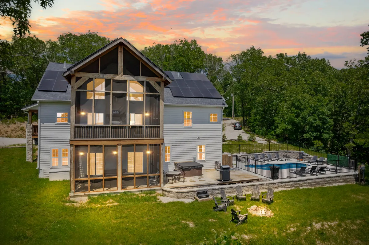 The image showcases a spacious, lodge-style home nestled among rolling Ozark hills, featuring a sparkling private pool, inviting fire pit, and expansive porch perfect for sunset views. Solar panels hint at eco-friendly amenities, while the tranquil setting is enhanced by lush surroundings and ample space for outdoor activities.