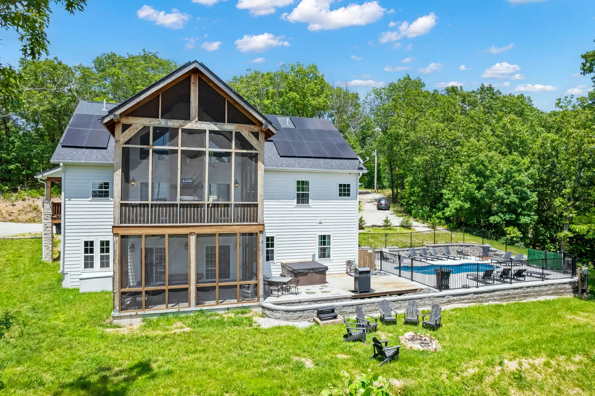 An inviting exterior scene featuring a screened porch, a sparkling swimming pool, a cozy fire pit, and lush greenery all around. The area is set up for swimming, barbecuing, and social gatherings, evoking a sense of summer relaxation and outdoor fun.