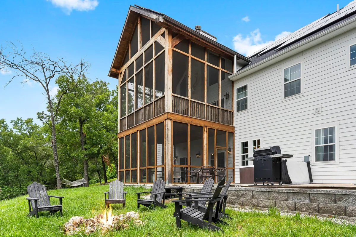 The image displays a spacious screened porch area featuring a fire pit surrounded by Adirondack chairs, a barbecue grill, and a lush green backdrop. The inviting setup suggests opportunities for outdoor gatherings, cozy evenings by the fire, and shared meals under the stars.