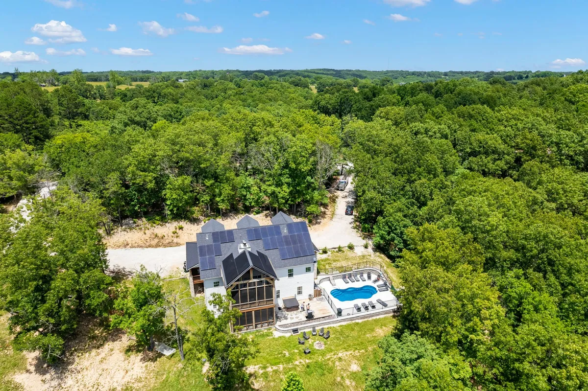 A sweeping exterior view showing the large home with solar panels, a sparkling swimming pool surrounded by trees, and inviting poolside seating. The driveway leads up to the home, and the setting is tranquil and immersed in nature.